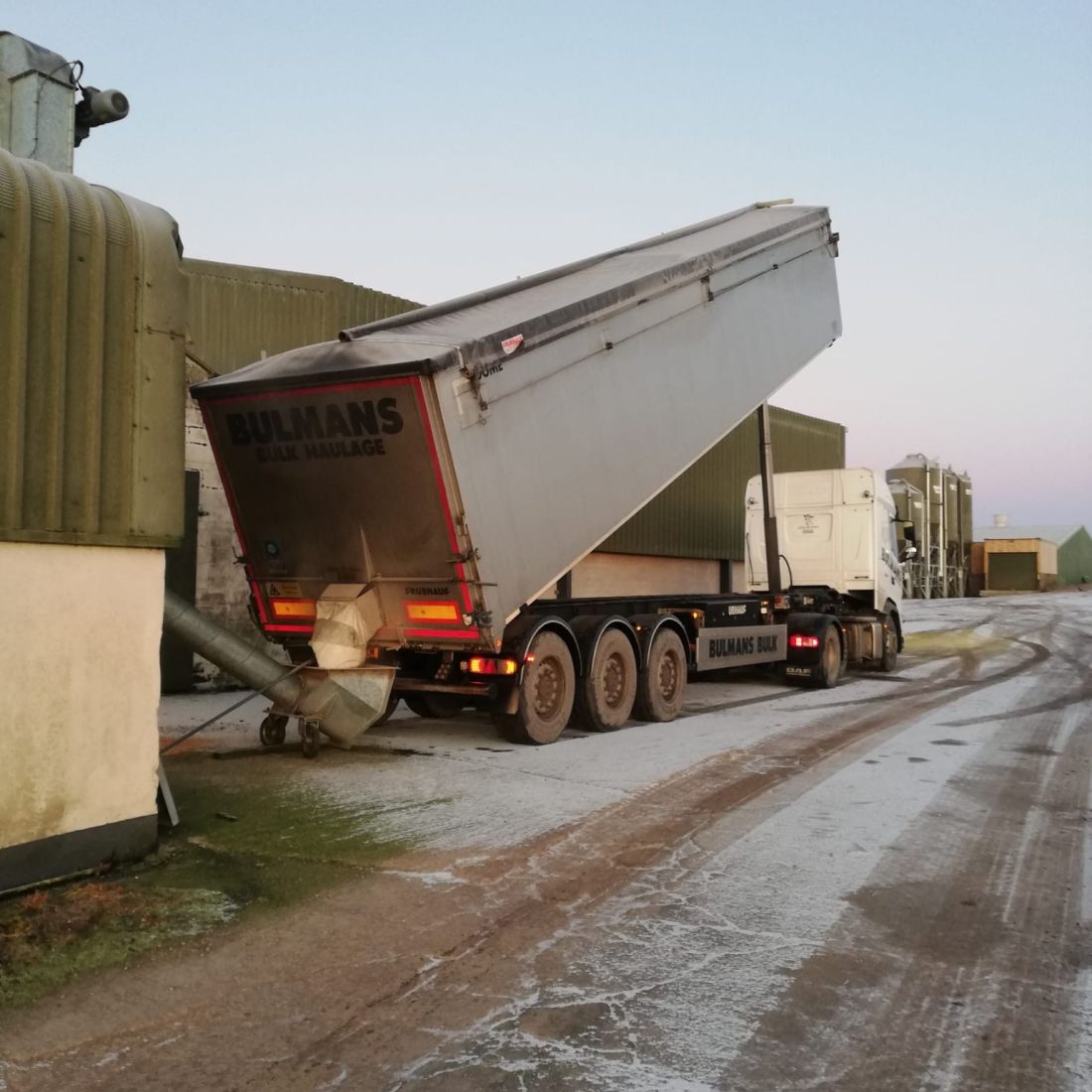 A Bulmans Bulk Haulage tipper truck unloads materials at a storage facility during dawn, on an icy day.