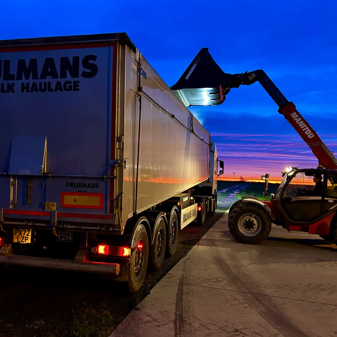 A Bulmans Bulk Haulage DAF Truck being loaded by a Manitou Articulated Loader, at dusk with a vivid yellow, purple and blue sunset in the background.