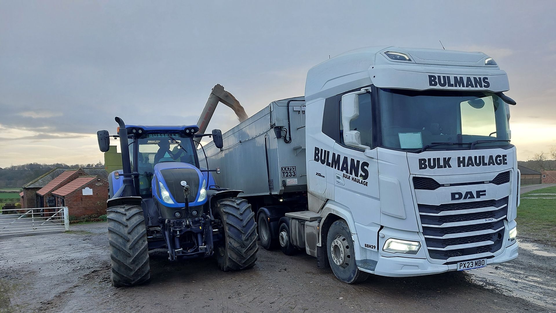A Bulmans Bulk Haulage DAF truck is being loaded by a blue tractor on a farm, illustrating the collaborative effort in agricultural bulk transport services.