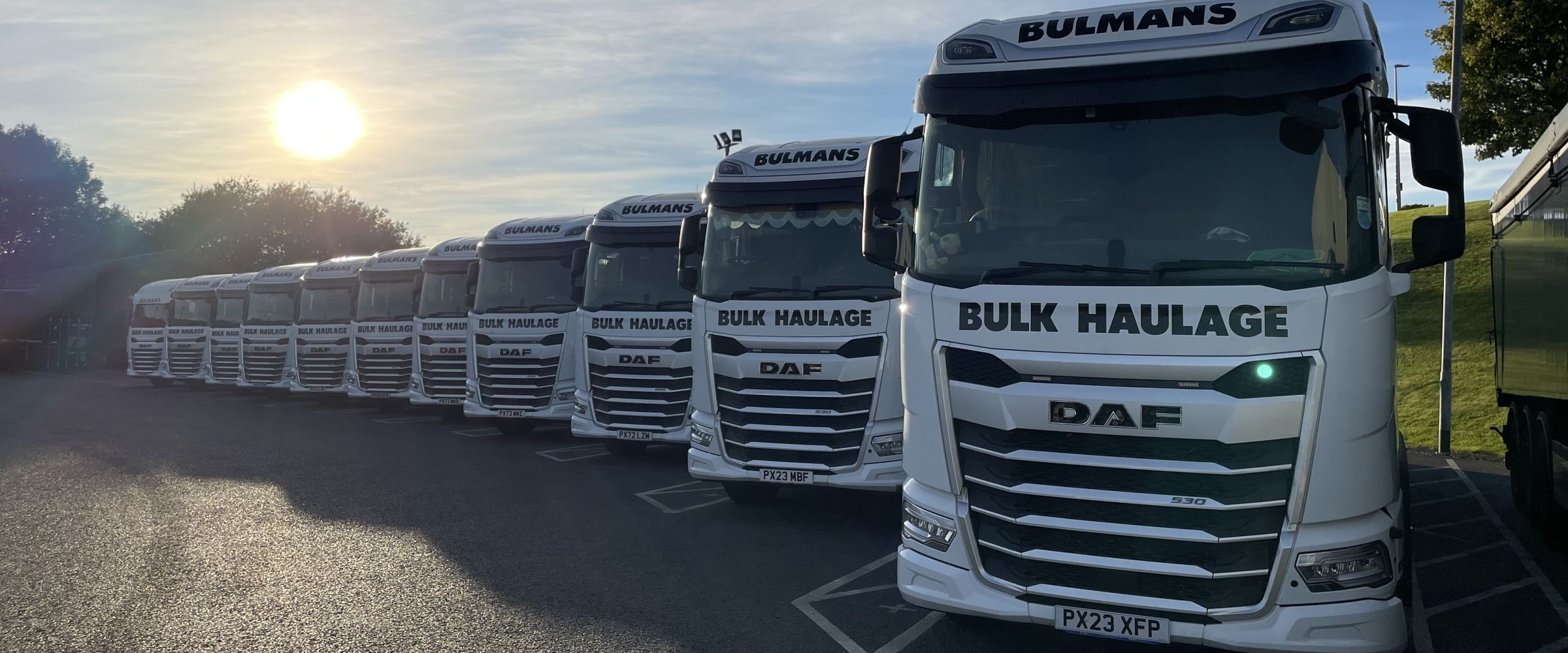 A row of Bulmans Bulk Haulage DAF white trucks parked in a line with a blue sky and setting sun above.