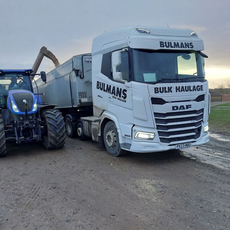 A Bulmans Bulk Haulage DAF truck is being loaded by a blue tractor on a farm, illustrating the collaborative effort in agricultural bulk transport services.